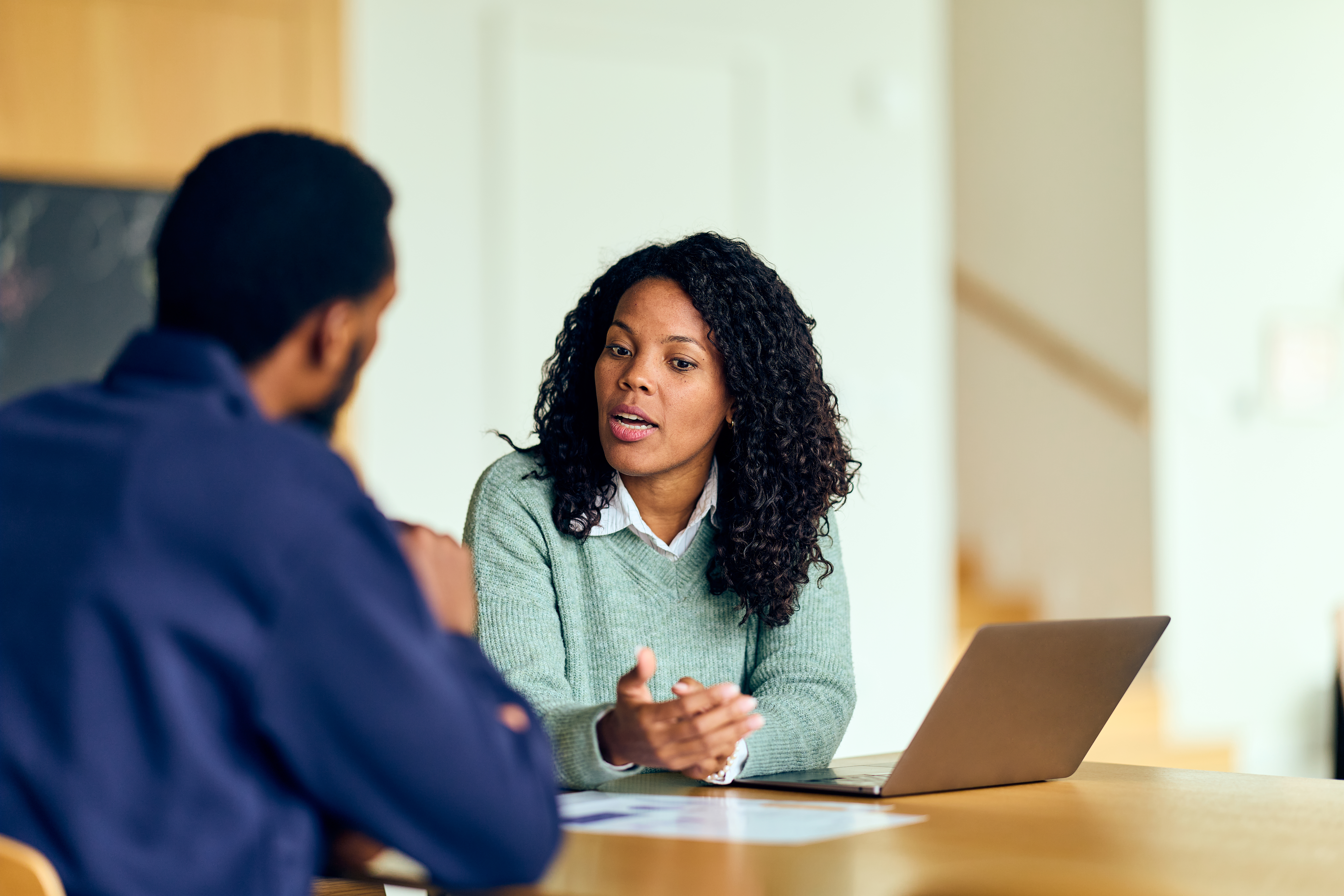 African-American woman leads business meeting with colleague, discussing ideas at a laptop-equipped office.